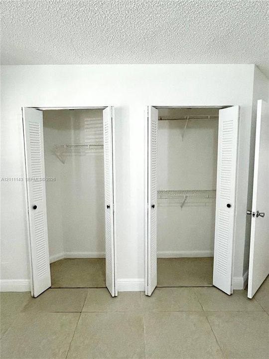 Dual reach-in closets featuring white louvered bi-fold doors, white interior shelving, and large-format neutral tile flooring