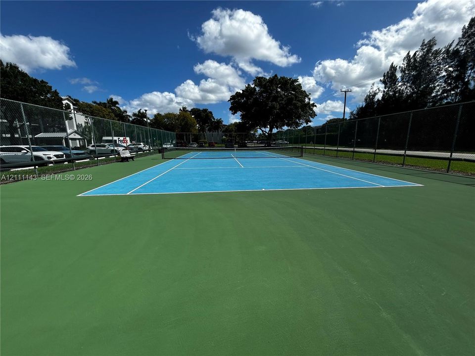 Tennis court featuring a high-contrast blue and green hard surface, full-perimeter chain-link fencing, and mature perimeter trees