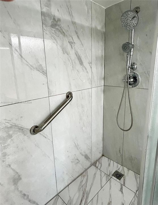 Shower enclosure featuring large-format veined tile walls and flooring, dual-function shower head with handheld attachment, chrome-finish fixtures, and a wall-mounted support bar