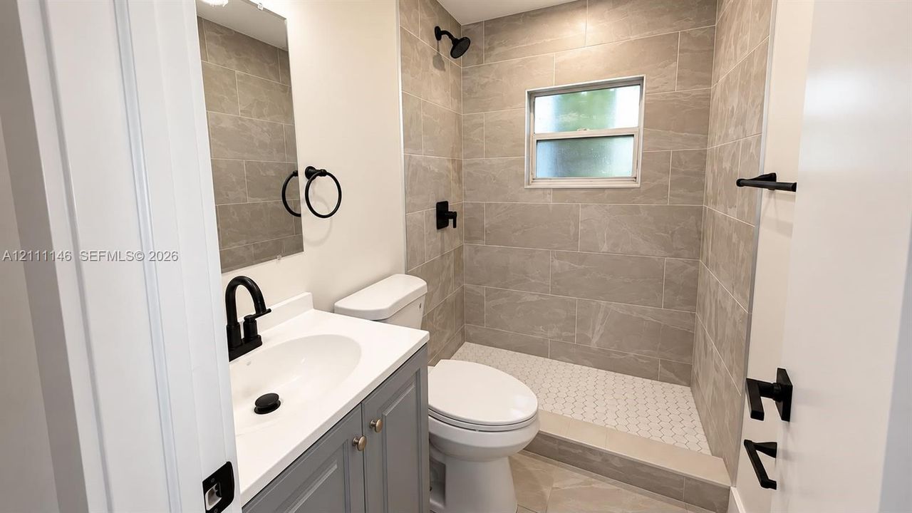 Bathroom featuring a grey vanity with a white countertop, matte black fixtures, walk-in shower with stone-tile surround, and hexagonal floor tile