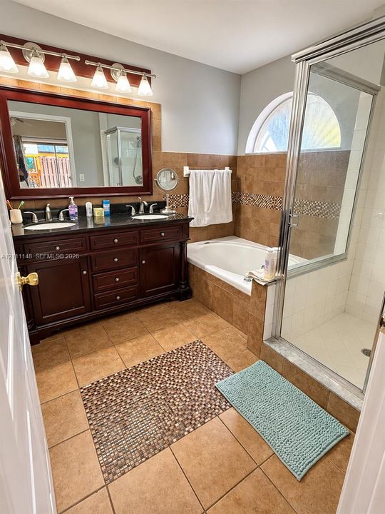 Dark wood double vanity with a stone countertop, built-in soaking tub, separate glass-enclosed shower, arched window, and multi-tone tile flooring