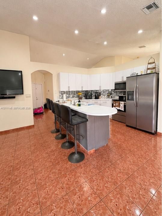 Kitchen featuring white cabinetry, solid-surface breakfast bar with integrated sink, stainless steel appliances, multi-toned decorative backsplash, and recessed lighting