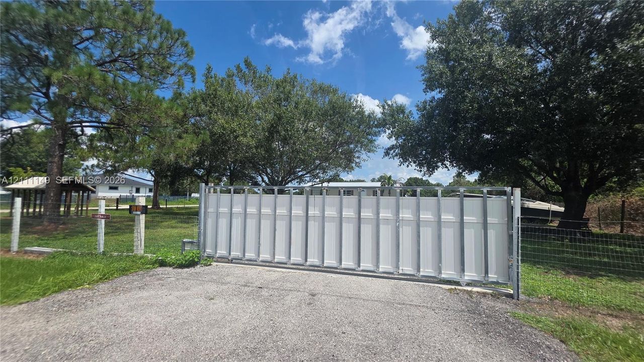 Metal sliding entry gate with white paneling, stone-aggregate driveway, perimeter wire fencing, and mature trees