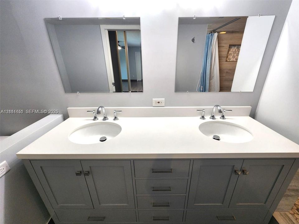 Double vanity featuring a light-toned solid surface countertop, dual undermount sinks, chrome widespread faucets, and gray Shaker-style cabinetry with matte finish hardware, topped by two frameless wall-mounted mirrors
