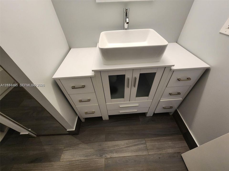 White vanity featuring a rectangular vessel sink, solid surface countertop, glass-panel cabinet doors, brushed metal hardware, and wood-finish flooring
