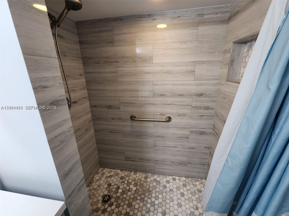 Walk-in shower with wood-finish tile wall surround, hexagonal floor tiling, recessed wall niche, stainless steel shower head, and wall-mounted grab bar