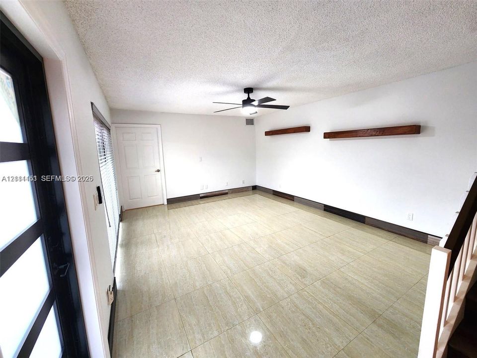 Neutral-toned tile flooring, dual wooden floating shelves, black-framed glass entry door, and a central ceiling fan with integrated light fixture