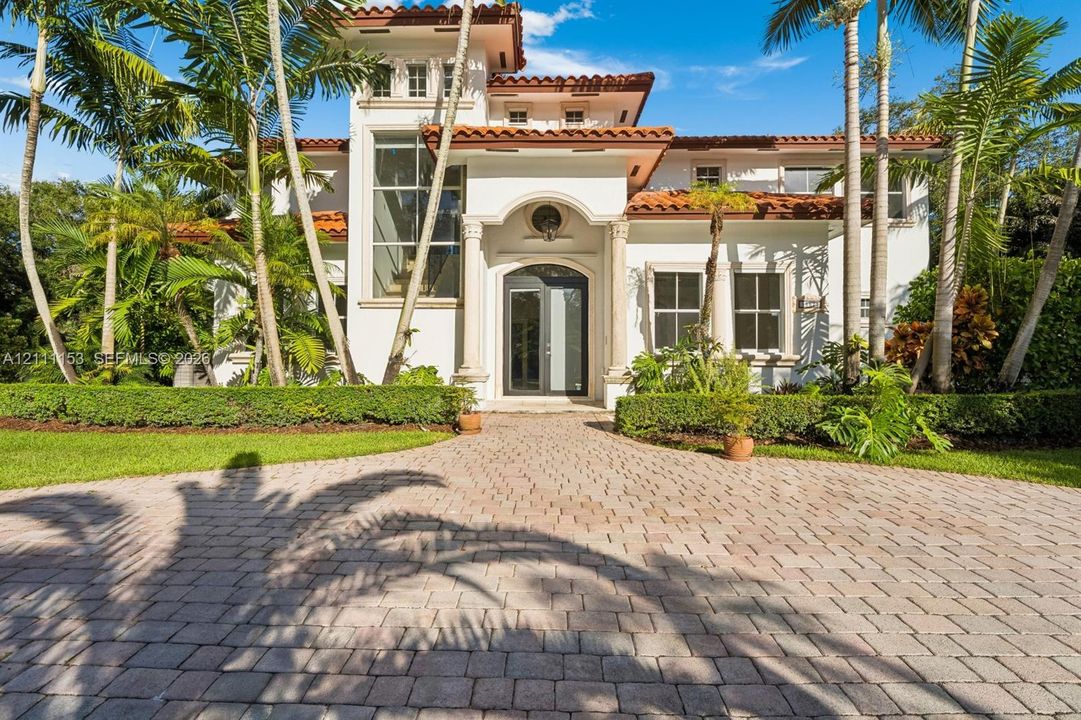 Mediterranean-inspired exterior featuring a clay tile roof, arched entryway with classical columns, stucco facade, multi-story glass window panel, and a paved brick driveway