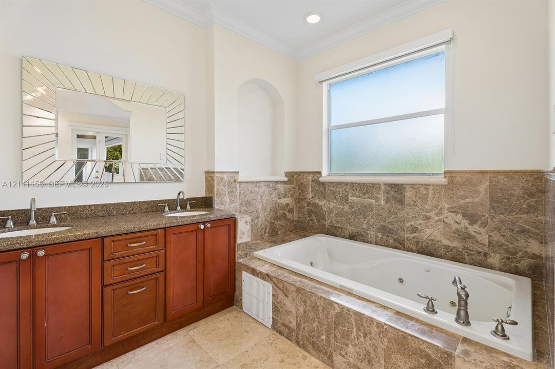 Dual-sink vanity with stone countertop, soaking tub with jetted features, full-height stone tile surround, built-in recessed wall niche, and decorative wall-mounted mirror