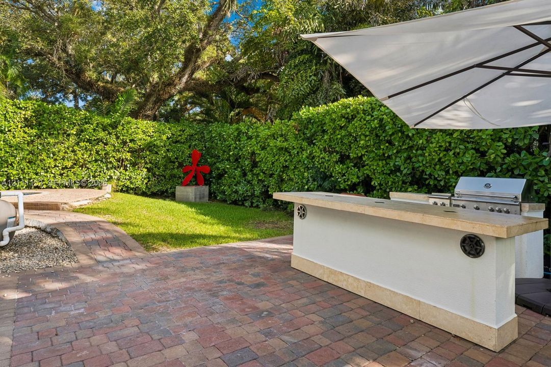 Outdoor kitchen featuring a built-in grill and stone-capped bar, set upon a red brick paver patio, surrounded by mature privacy hedging and a manicured lawn