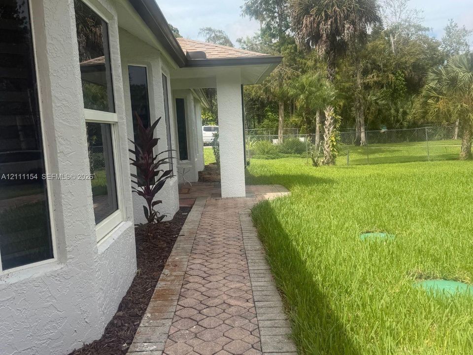 Stucco exterior with a paved stone walkway, overhanging roofline, and expansive lawn with perimeter fencing