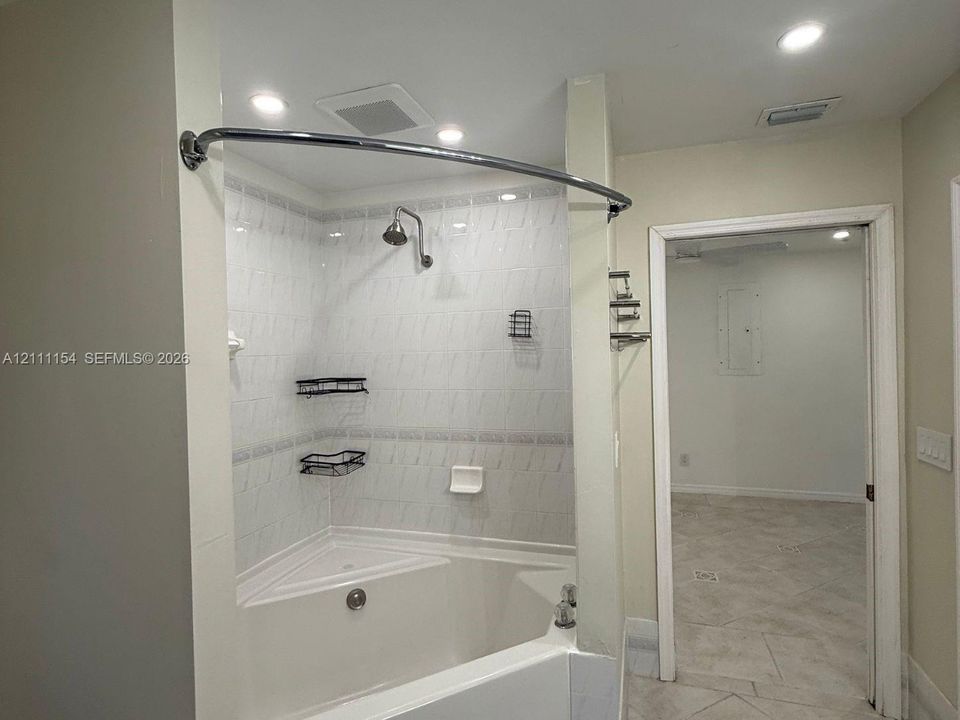 Corner soaking tub with integrated shower head, white tiled surround with decorative inlay, curved shower rod, and recessed ceiling lighting
