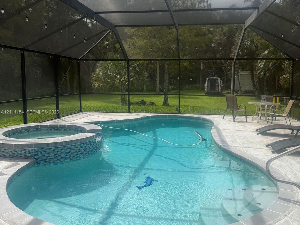 Screen-enclosed pool area featuring an integrated spa with decorative tile accents, stone-finish decking, and string lighting