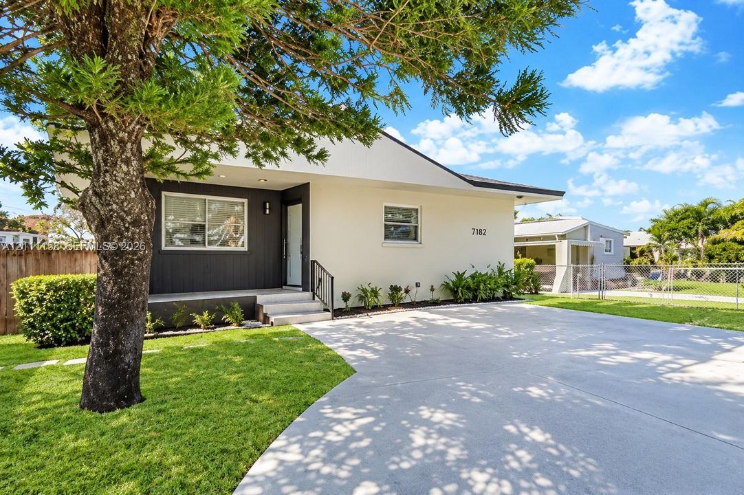 Exterior elevation featuring a light-toned stucco facade with dark vertical siding accents, an expansive concrete driveway, a covered front entryway, and mature landscaping
