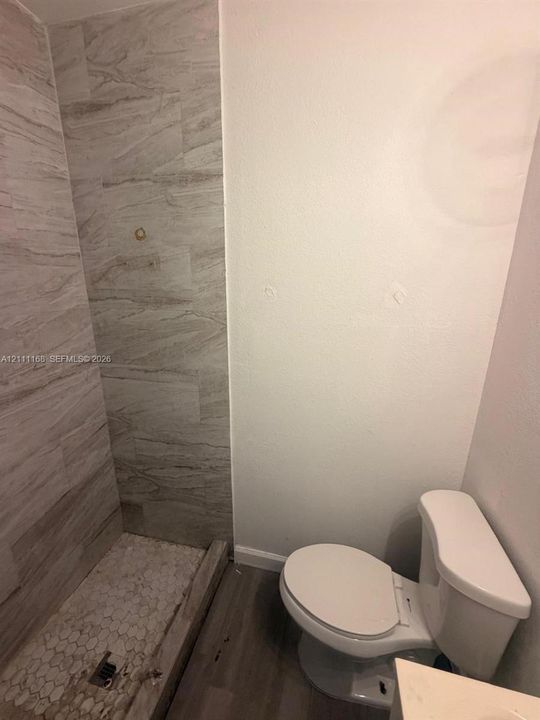 Walk-in shower featuring large-format neutral tile wall surrounds and a decorative hexagonal tile floor, paired with a low-profile toilet and wood-finish flooring