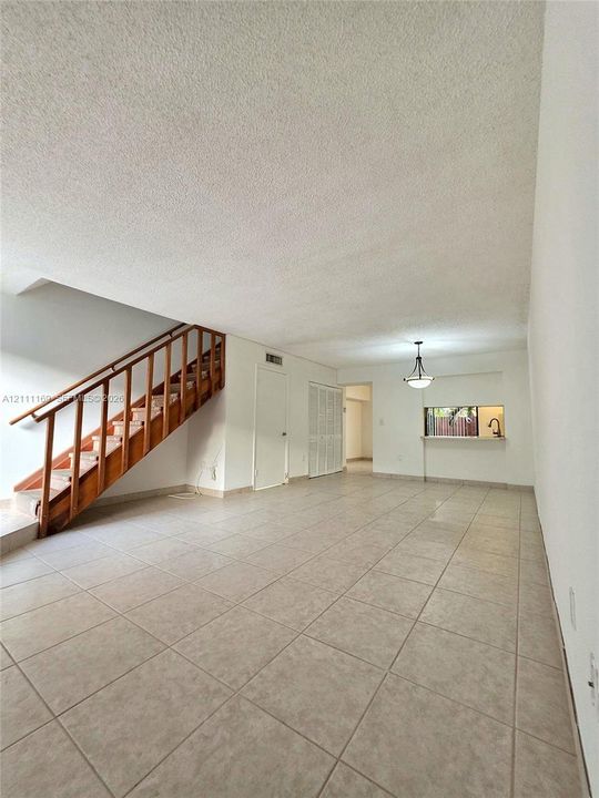 Open floor plan featuring neutral tile flooring, a staircase with a wooden handrail and patterned stair runner, and a pass-through opening with a pendant light fixture