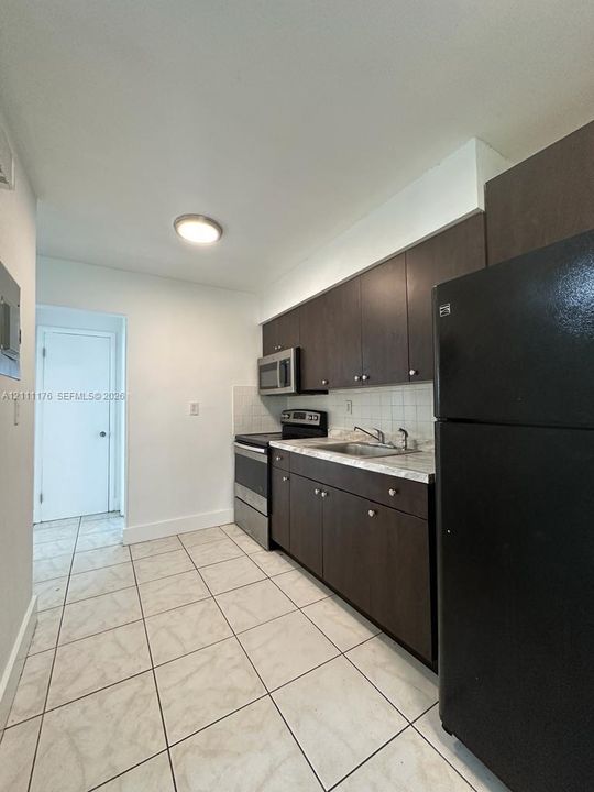 Kitchen featuring dark-finish cabinetry, stone-patterned countertops, stainless steel appliances, ceramic tile flooring, and a tiled backsplash