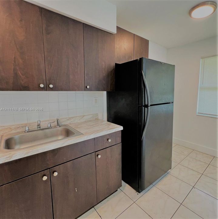 Dark-finish cabinetry, stone-pattern laminate countertops, stainless steel drop-in sink, black full-size refrigerator, and ceramic tile flooring
