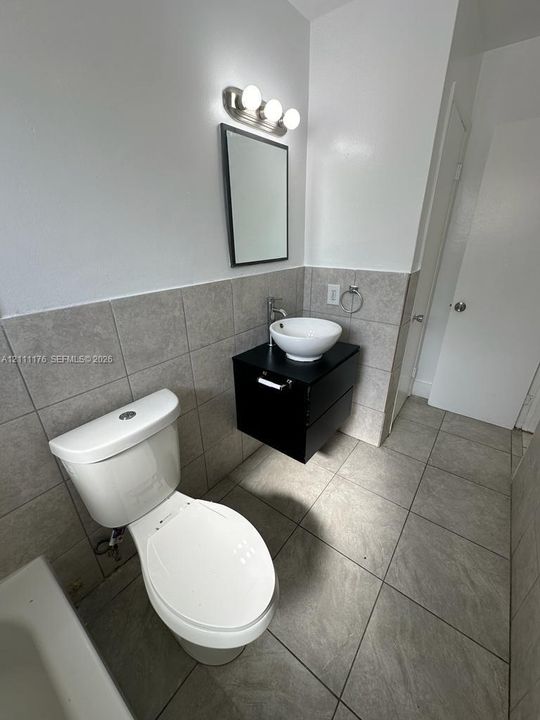 Floating vanity with a circular vessel sink, wall-mounted faucet, neutral large-format tile flooring, coordinating tiled wall surround, and three-bulb vanity lighting
