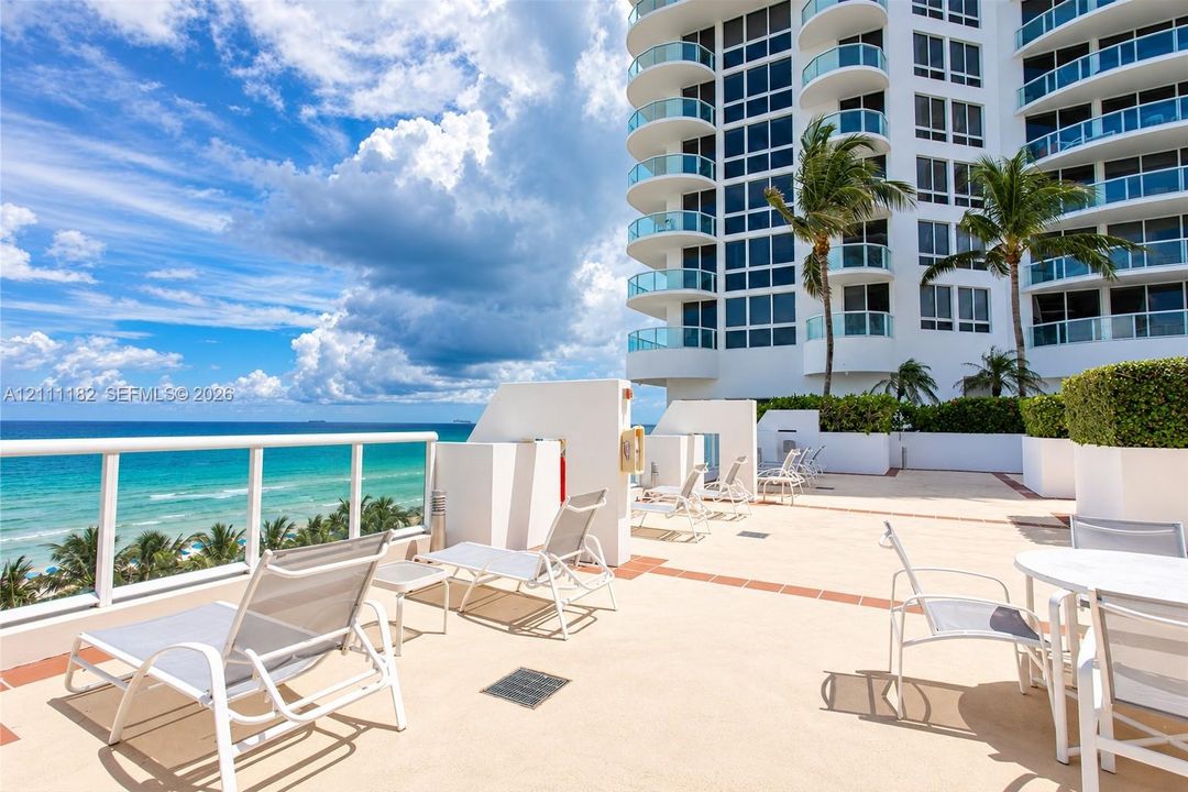 Expansive outdoor terrace featuring stone-finish flooring, glass-paneled railings, high-rise architectural facade, and manicured perimeter hedges