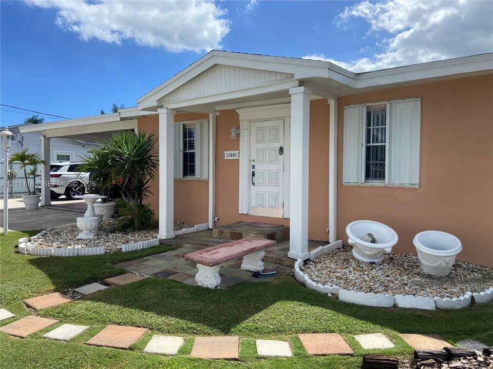 Covered front porch featuring square columns, decorative paneled entrance door, stone-tiled patio, and manicured landscaping with stone-accented garden beds