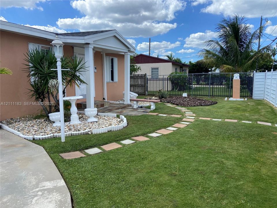 Exterior space featuring a manicured lawn, stone pathway, rock garden beds, and perimeter metal fencing
