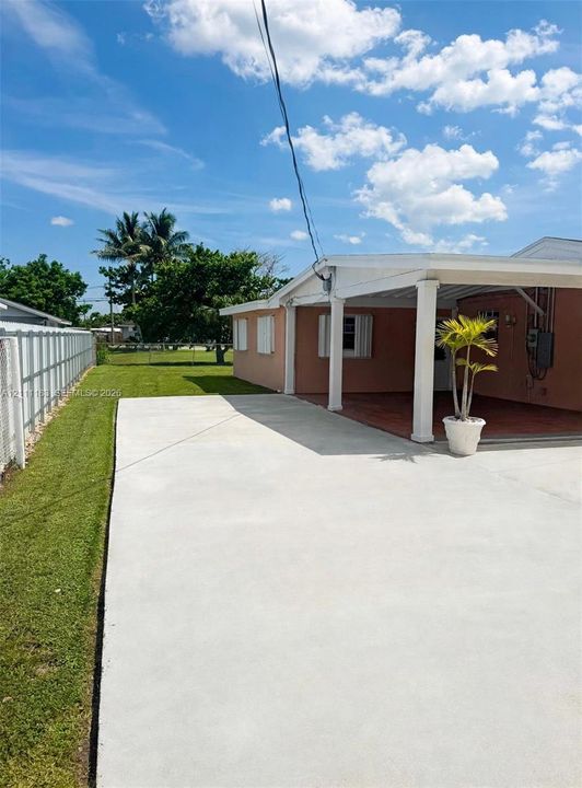 Exterior area featuring a paved concrete driveway, covered patio, manicured lawn, and perimeter fencing
