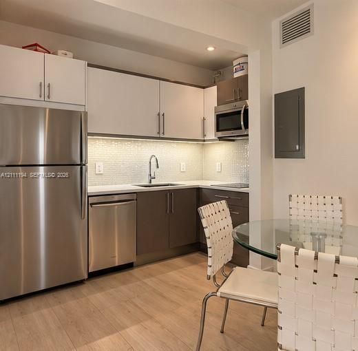 Kitchen featuring stainless steel appliances, two-tone flat-panel cabinetry, light-toned solid surface countertops, mosaic tile backsplash, and wood-finish flooring