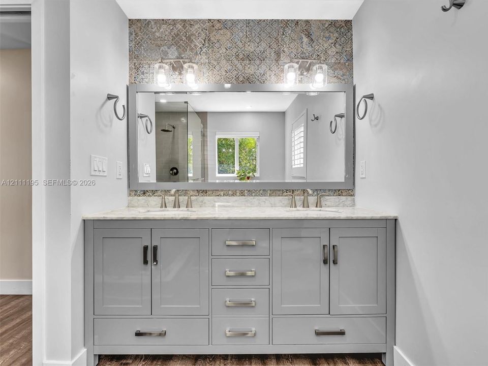 Dual-sink vanity featuring a light-toned stone countertop, grey shaker-style cabinetry, a large rectangular mirror, patterned tile backsplash, and dual-light fixtures