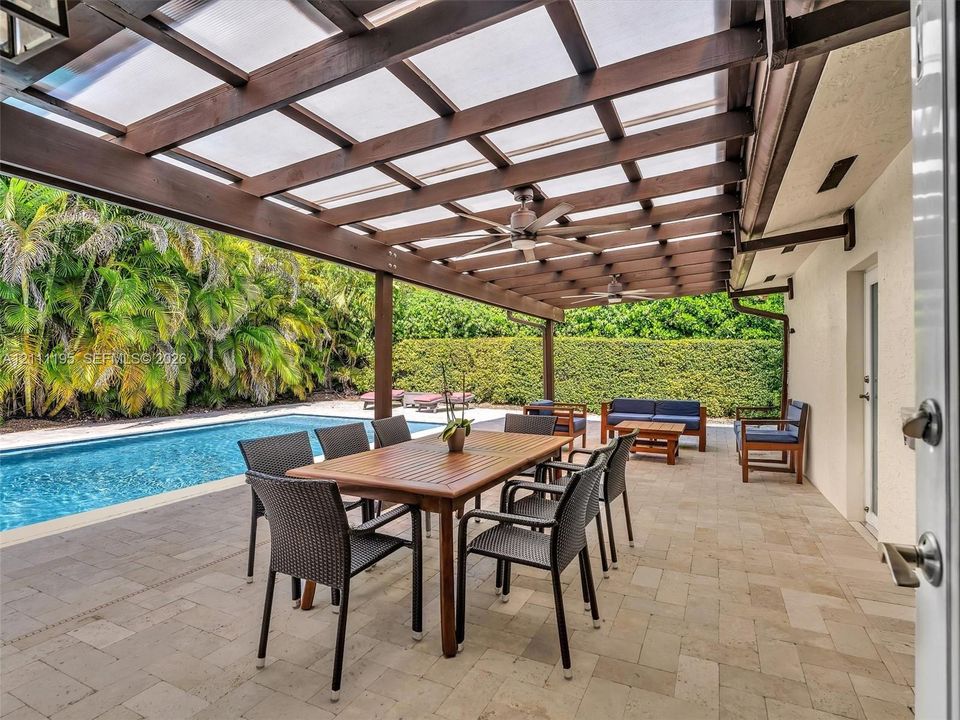 Covered patio featuring a timber-frame pergola with translucent roofing panels and ceiling fans, stone-paver flooring, and a rectangular swimming pool