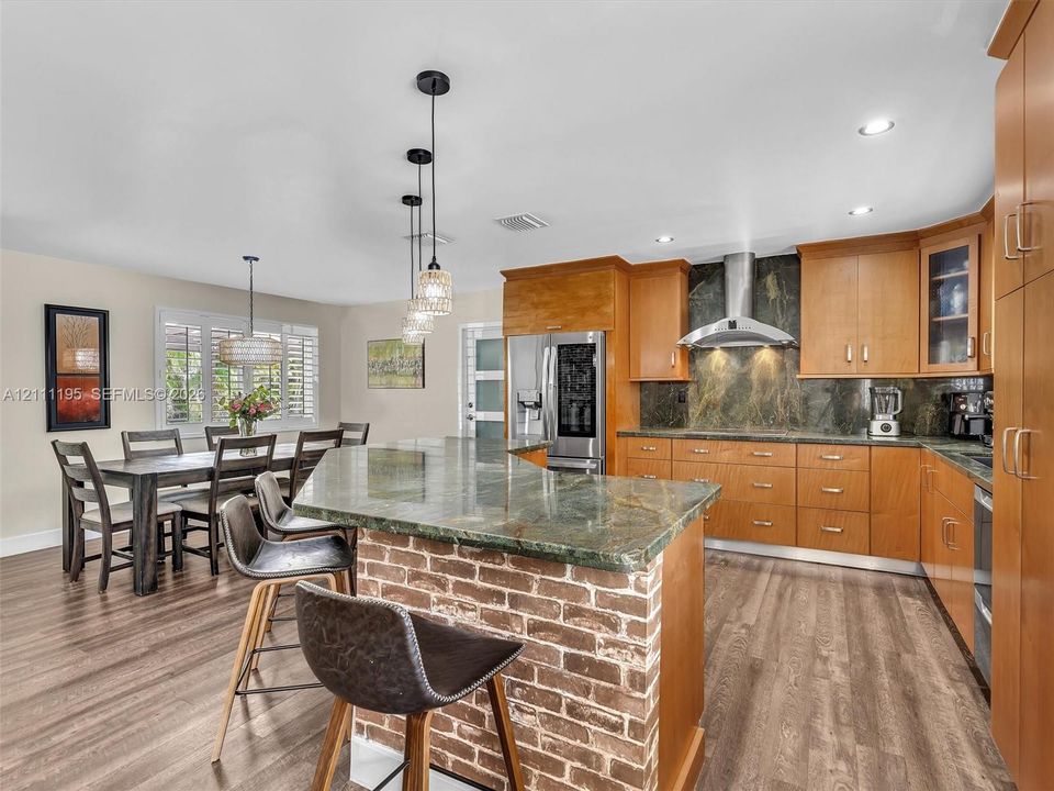 Kitchen featuring wood cabinetry, an island with a brick base and stone countertop, stone backsplash, stainless steel range hood, and wood-finish flooring
