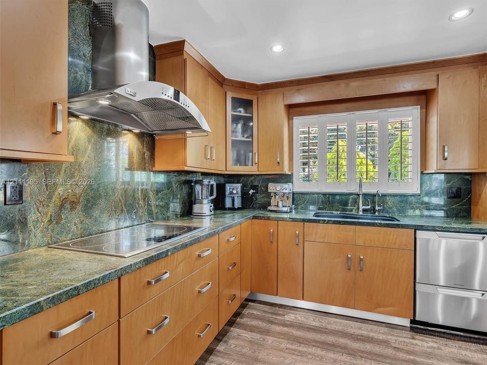 Custom wood-veneer cabinetry, stone countertops with matching full-height backsplash, stainless steel range hood, integrated stainless steel dishwasher, and shutter-style window coverings