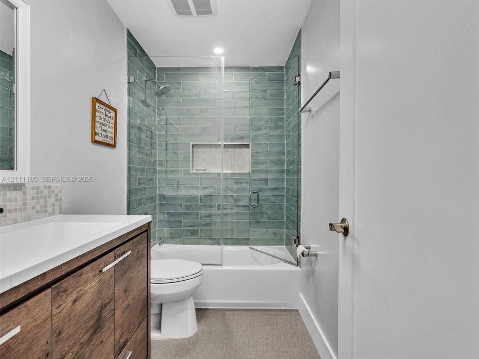Bathroom featuring a wood-finish vanity with a rectangular sink, full-height blue-green subway tile shower surround, integrated wall niche, and glass panel door