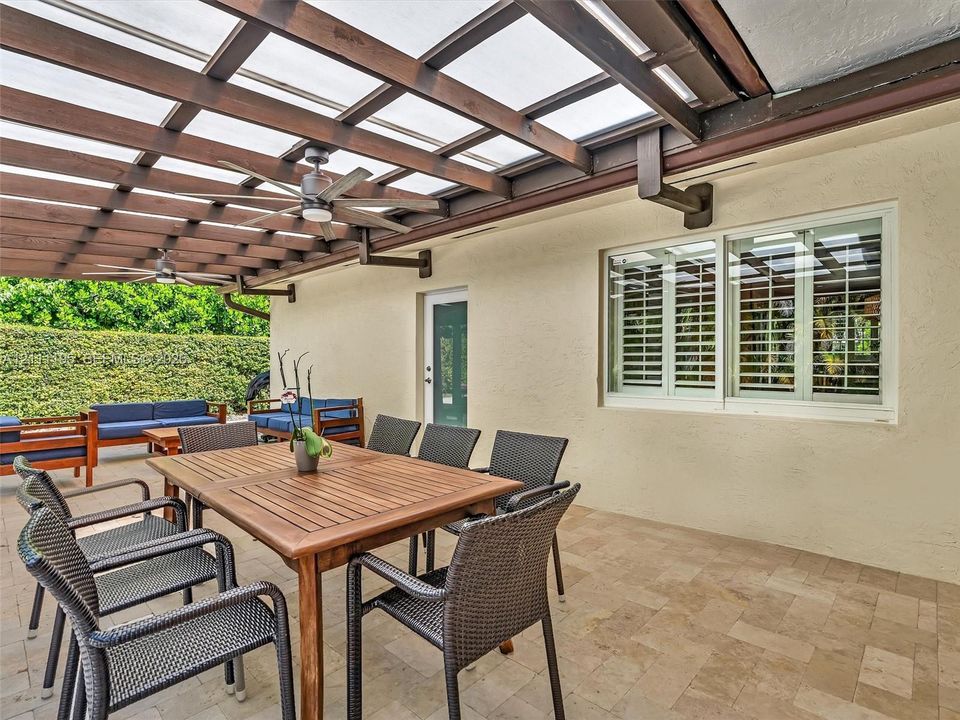 Covered patio featuring a timber-frame pergola, stone paver flooring, dual ceiling fans, and windows equipped with plantation shutters