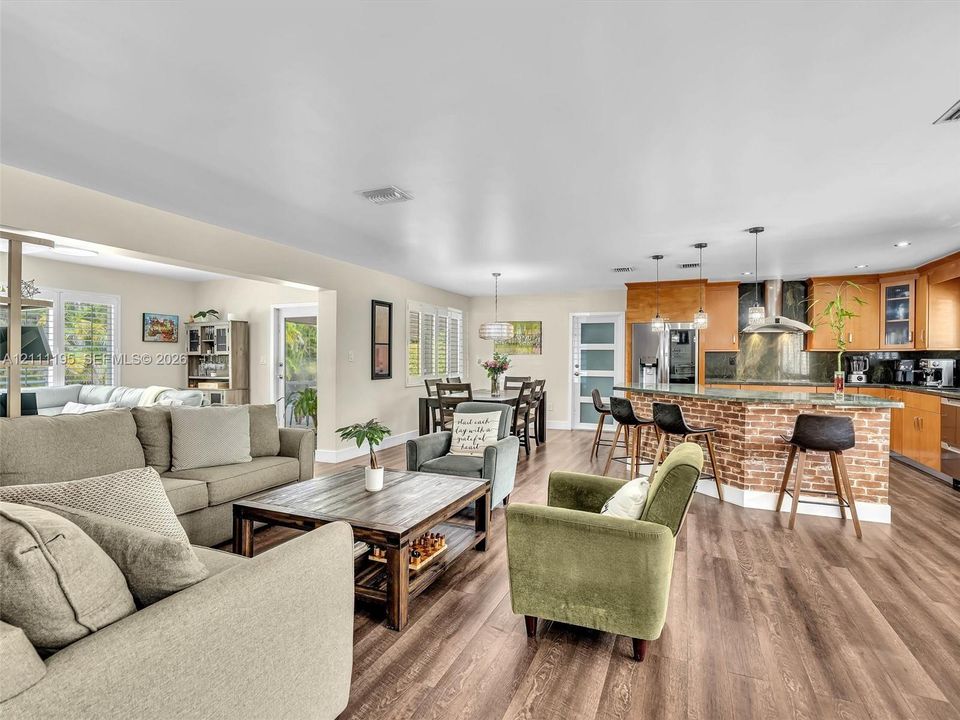 Open-concept interior featuring wood-finish flooring, kitchen island with brick-veneer base, stainless steel range hood, pendant lighting, and interior shutters