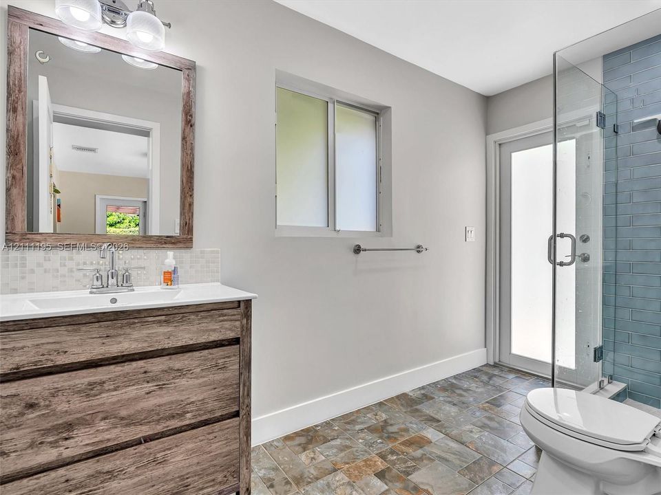 Bathroom featuring a wood-finish vanity with an integrated basin, a framed mirror, natural stone-finish floor tiling, and a walk-in shower with blue tile surround and a frameless glass door