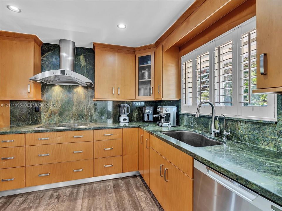 Kitchen featuring natural stone countertops and full-height backsplash, wood-finish cabinetry, stainless steel range hood, undermount sink with gooseneck faucet, and plantation shutter window treatments