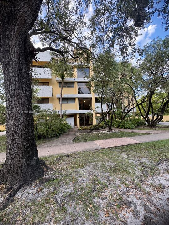Multi-story building exterior featuring concrete construction, stackable balconies, large glass windows, and mature shade trees