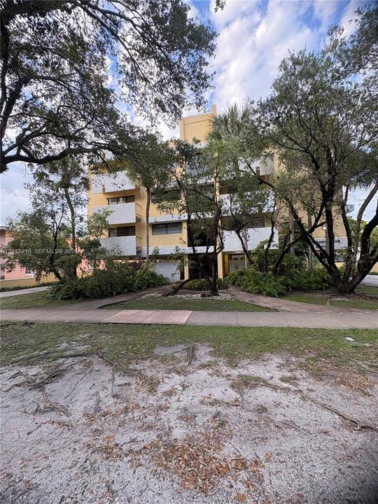 Multi-story building exterior featuring concrete construction, white balcony railings, and mature canopy trees