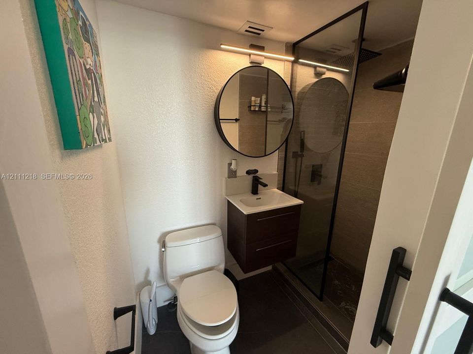 Modern floating vanity with a light-toned countertop and matte black faucet, paired with a circular mirror, linear vanity lighting, and a glass-enclosed shower with tiled surround
