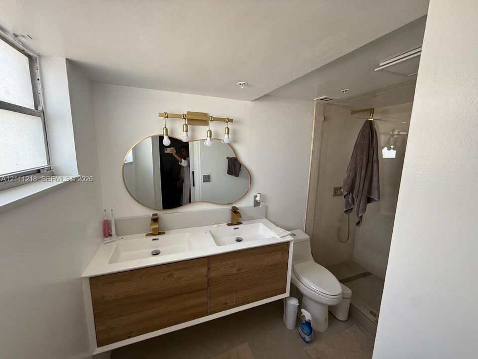 Floating dual-sink vanity with wood-finish cabinetry, quartz countertop, brass-toned fixtures, organic-shaped mirror, and glass-enclosed shower