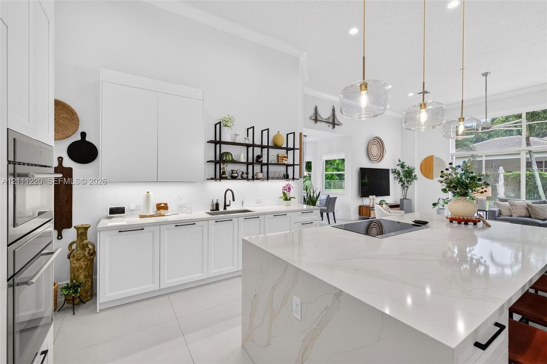 Kitchen featuring a large central island with a stone-surface countertop, high-gloss white cabinetry, built-in stainless steel double wall ovens, matte black hardware, and glass pendant lighting