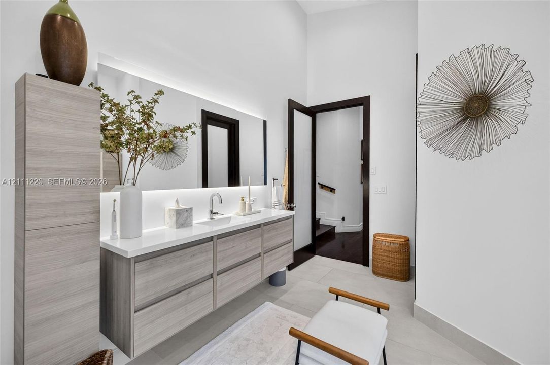 Floating vanity with wood-finish cabinetry, solid-surface countertop, integrated rectangular sink, wall-to-wall mirror with integrated LED backlighting, and floor-to-ceiling cabinetry