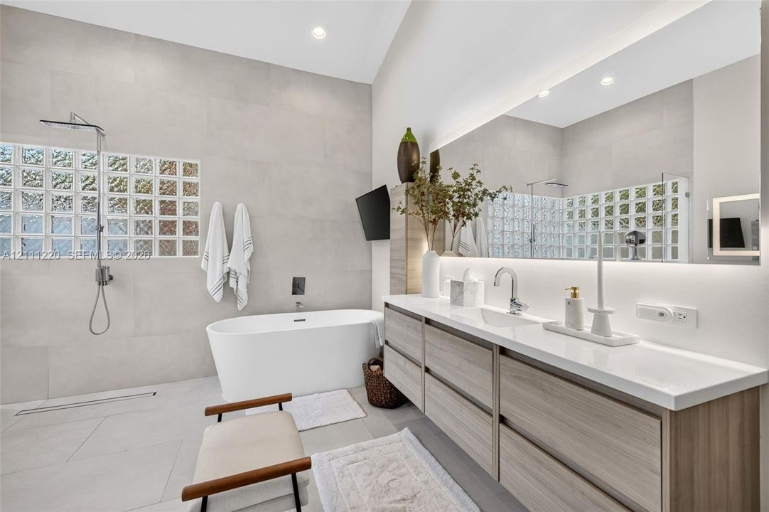 Bathroom featuring a floating wood-finish vanity with a countertop basin, a standalone soaking tub, full-wall tiling, a glass-block privacy window, and an expansive backlit mirror
