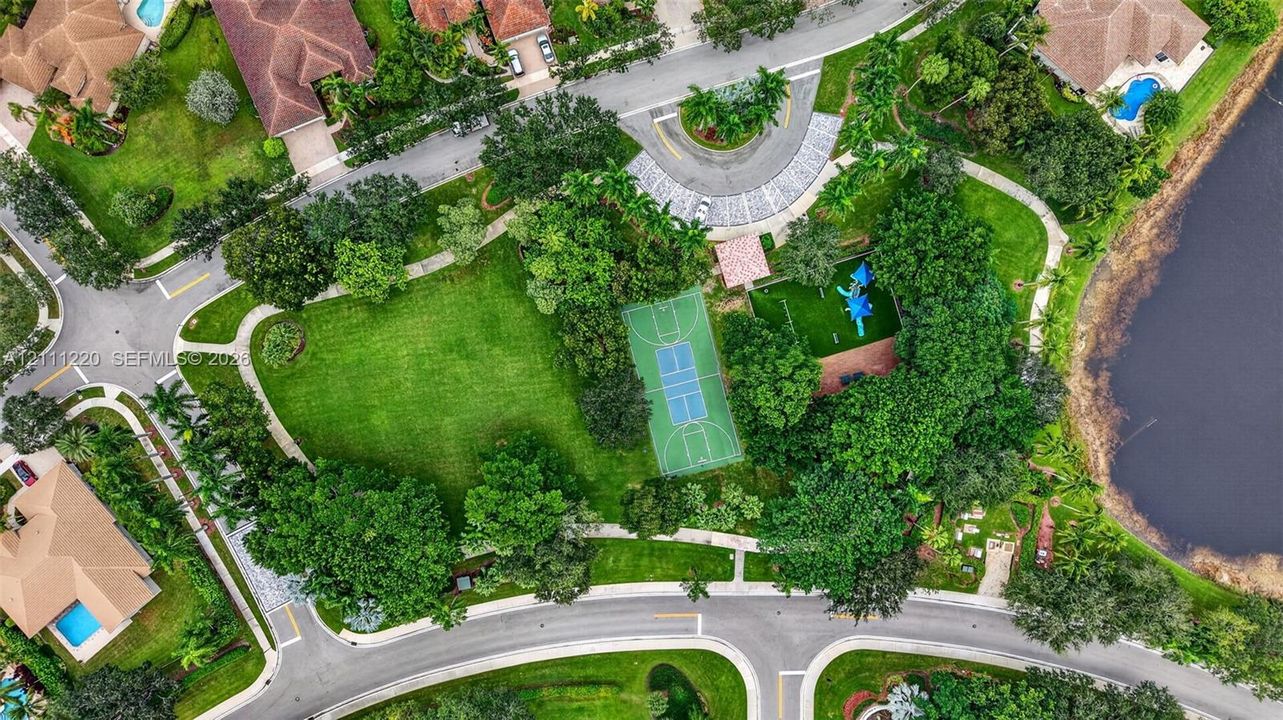 Aerial perspective of a community recreation area featuring a basketball court, playground, expansive green space, mature trees, and a bordering water feature
