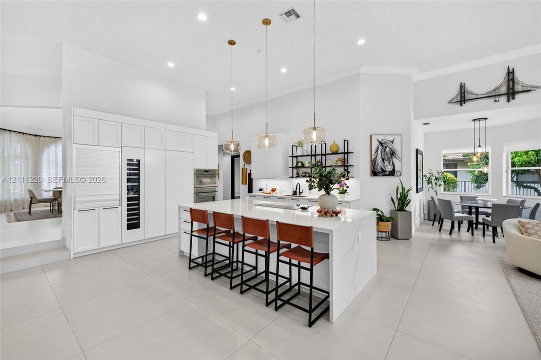 Gourmet kitchen featuring a large central island with stone-finish countertops, custom floor-to-ceiling white cabinetry, integrated stainless steel appliances, tiered pendant lighting, and oversized neutral tile flooring