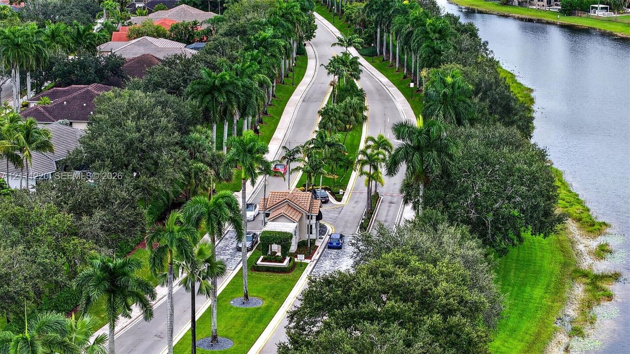 Gated entrance featuring a tile-roof structure, bordered by palm-lined streets, mature landscaping, and an expansive waterway