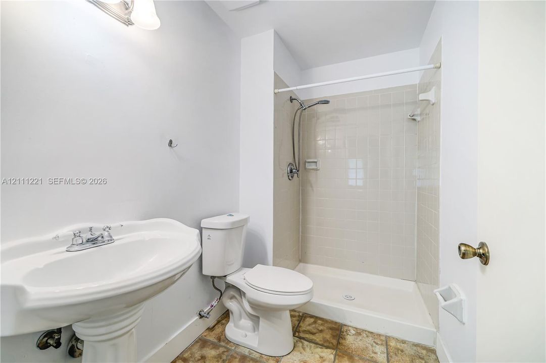 Wall-mounted pedestal sink, ceramic tile shower surround, shower head with integrated handheld sprayer, and stone-finish tile flooring