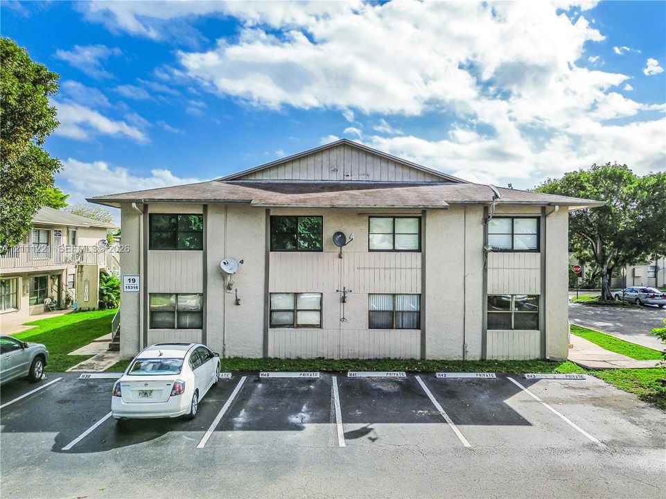 Two-story exterior featuring light-toned stucco siding, a low-pitch roofline, rectangular windows, and dedicated asphalt surface parking