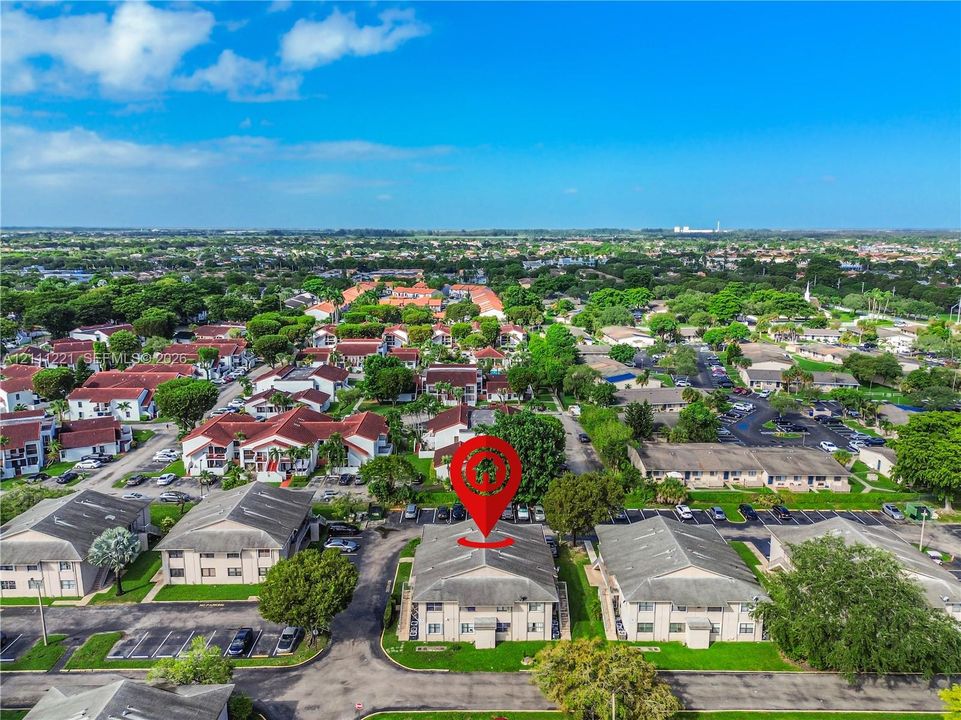 Aerial perspective of a multi-story residential building featuring a light-toned exterior, gabled rooflines, designated surface parking, and manicured green space